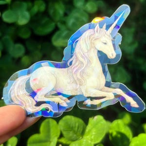 May include: A holographic sticker featuring a white unicorn with a flowing mane and a long horn. The unicorn is in a reclining pose, with a blue geometric border. The sticker is held against a backdrop of green leaves.