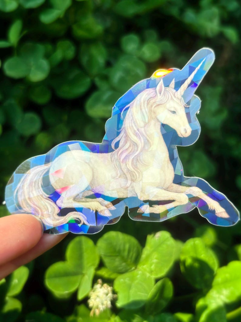 May include: A holographic sticker featuring a white unicorn with a flowing mane and a long horn. The unicorn is in a reclining pose, with a blue geometric border. The sticker is held against a backdrop of green leaves.