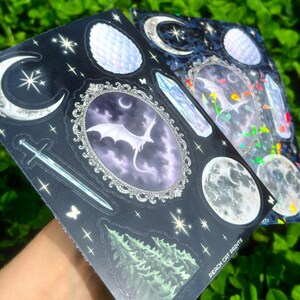 May include: Two sticker sheets with a dark background, featuring celestial and fantasy designs. One sheet has a dragon in an oval frame, moons, stars, and a sword. The other sheet has similar designs with iridescent accents. The text "PEACH CAT PRINTS" is visible.