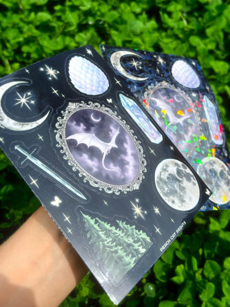 May include: Two sticker sheets with a dark background, featuring celestial and fantasy designs. One sheet has a dragon in an oval frame, moons, stars, and a sword. The other sheet has similar designs with iridescent accents. The text "PEACH CAT PRINTS" is visible.