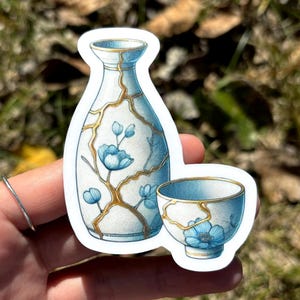 May include: A sticker depicting a white and blue vase and cup with gold detailing and blue floral patterns. The vase and cup are styled in the Kintsugi method, with gold lines filling the cracks. The sticker is on a white background.