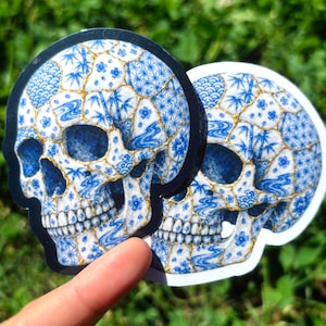 May include: Two skull-shaped stickers featuring a blue and white porcelain pattern with gold-colored cracks. The design includes various motifs such as bamboo, cherry blossoms, and waves, mimicking the appearance of kintsugi-repaired ceramic pottery.