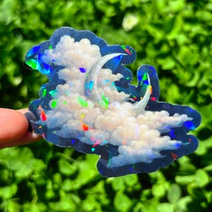May include: Holographic sticker featuring a crescent moon and fluffy clouds. The sticker has a dark blue border and iridescent accents that reflect multiple colors. The design is reminiscent of a dreamy night sky.