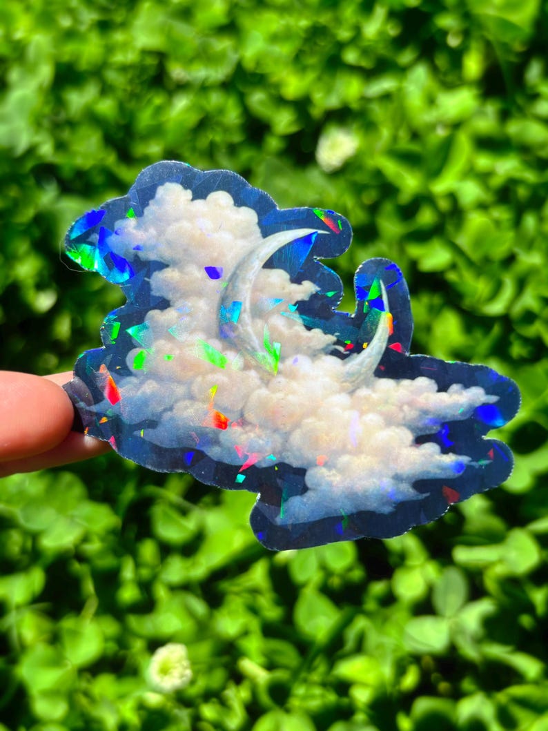 May include: Holographic sticker featuring a crescent moon and fluffy clouds. The sticker has a dark blue border and iridescent accents that reflect multiple colors. The design is reminiscent of a dreamy night sky.