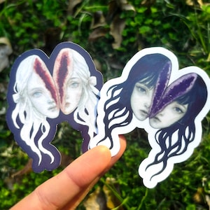 May include: Two stickers featuring a split-face design with a surreal, artistic style. One sticker has a white-haired woman with a red split, the other has a black-haired woman with a purple split. The stickers are held against a green, grassy background.