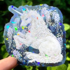 Puede incluir: Una pegatina hologr&aacute;fica con un unicornio blanco con una larga melena y un cuerno en espiral. El unicornio est&aacute; rodeado de elementos florales y descansa sobre un fondo azul oscuro con acentos iridiscentes. Dise&ntilde;o art&iacute;stico &uacute;nico.