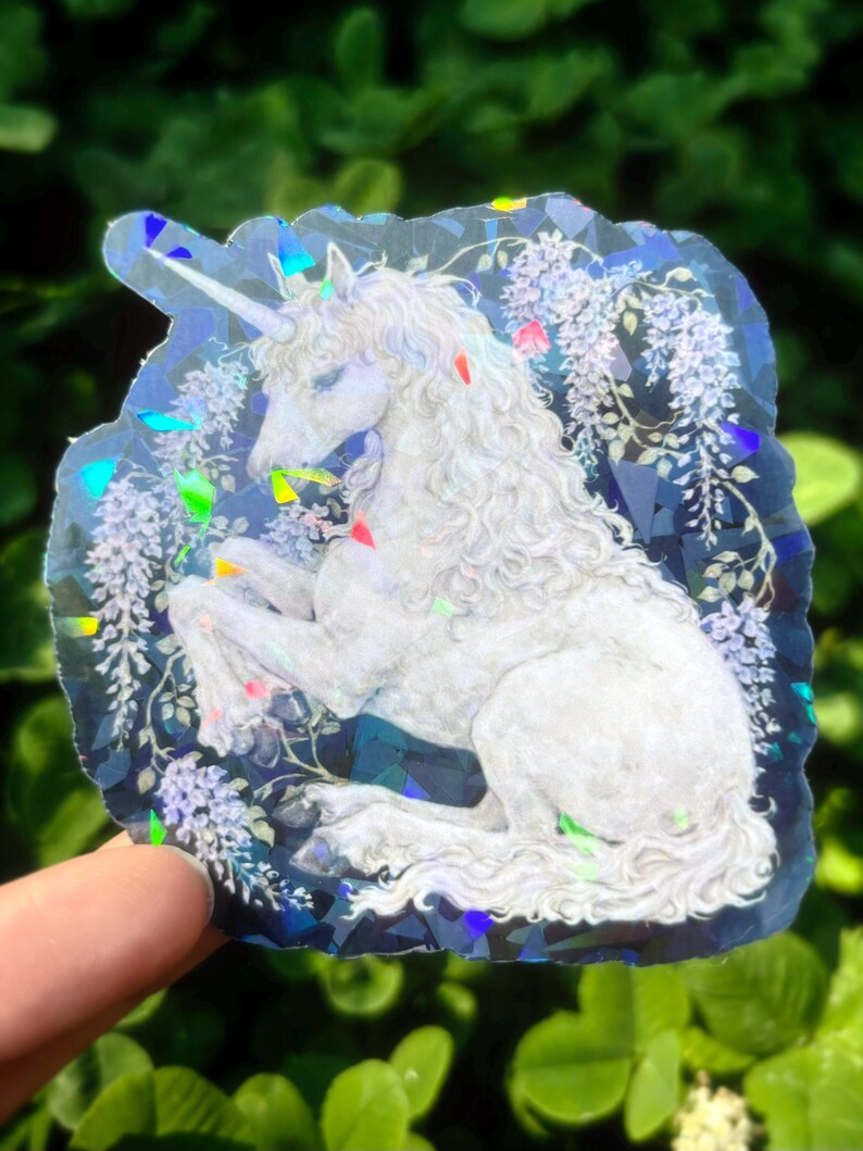 Puede incluir: Una pegatina hologr&aacute;fica con un unicornio blanco con una larga melena y un cuerno en espiral. El unicornio est&aacute; rodeado de elementos florales y descansa sobre un fondo azul oscuro con acentos iridiscentes. Dise&ntilde;o art&iacute;stico &uacute;nico.