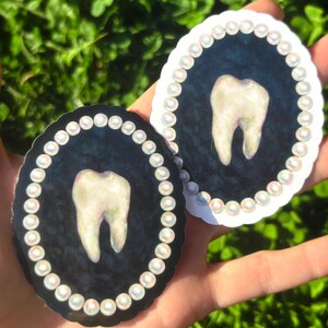 May include: Two oval stickers with a tooth design. One has a black background, the other a white background. Both have a pearl border and a tooth illustration in the centre. The stickers are held in a hand.