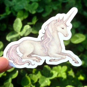 May include: A sticker featuring a watercolor illustration of a white unicorn with a long, flowing mane and tail. The unicorn is depicted in a resting pose, with its horn pointing upwards. The sticker has a white border and is set against a background of green foliage.