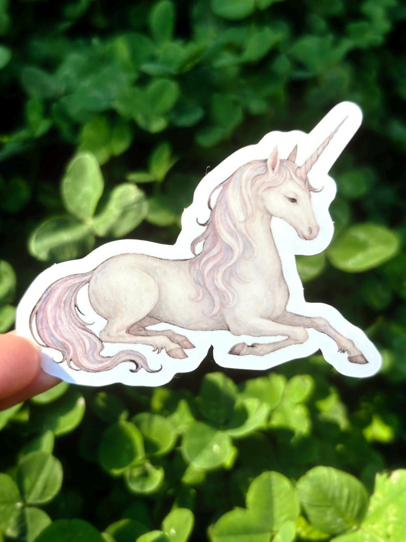 May include: A sticker featuring a watercolor illustration of a white unicorn with a long, flowing mane and tail. The unicorn is depicted in a resting pose, with its horn pointing upwards. The sticker has a white border and is set against a background of green foliage.