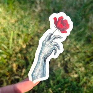 May include: A sticker featuring a detailed illustration of a metallic gauntlet holding a vibrant red rose. The gauntlet is rendered in shades of gray and silver, with intricate armor detailing. The rose has lush red petals. The sticker is held against a backdrop of green grass.