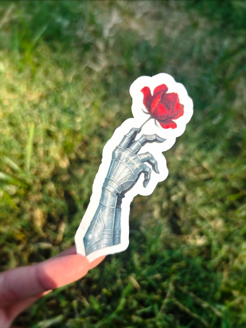 May include: A sticker featuring a detailed illustration of a metallic gauntlet holding a vibrant red rose. The gauntlet is rendered in shades of gray and silver, with intricate armor detailing. The rose has lush red petals. The sticker is held against a backdrop of green grass.