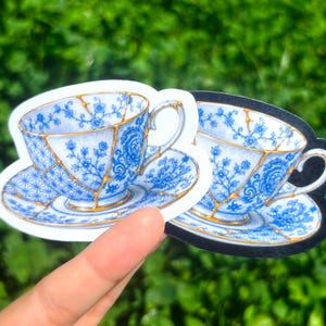 May include: Two die-cut stickers featuring a blue and white floral patterned teacup and saucer. The design includes gold-colored lines resembling kintsugi repair, with one sticker having a white border and the other having a black border.