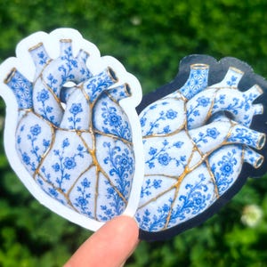 May include: Two anatomical heart stickers featuring a blue and white floral porcelain pattern with gold-colored cracks running across the surface. One sticker has a white border, while the other has a black border, both depicting the same design.