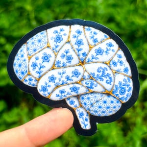 May include: A die-cut sticker shaped like a human brain, featuring a white surface decorated with blue floral patterns and geometric designs. The sections are separated by gold-colored lines, mimicking the appearance of kintsugi pottery.