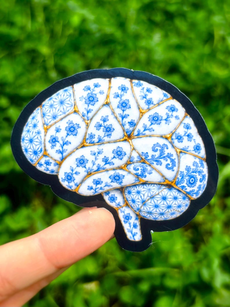 May include: A die-cut sticker shaped like a human brain, featuring a white surface decorated with blue floral patterns and geometric designs. The sections are separated by gold-colored lines, mimicking the appearance of kintsugi pottery.