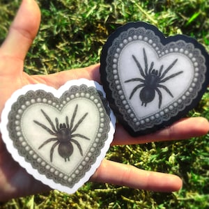 May include: Two heart-shaped stickers with a black spider design. The spiders are on a white background, framed by a grey lace border with pearl accents. The stickers are held against a backdrop of green grass.