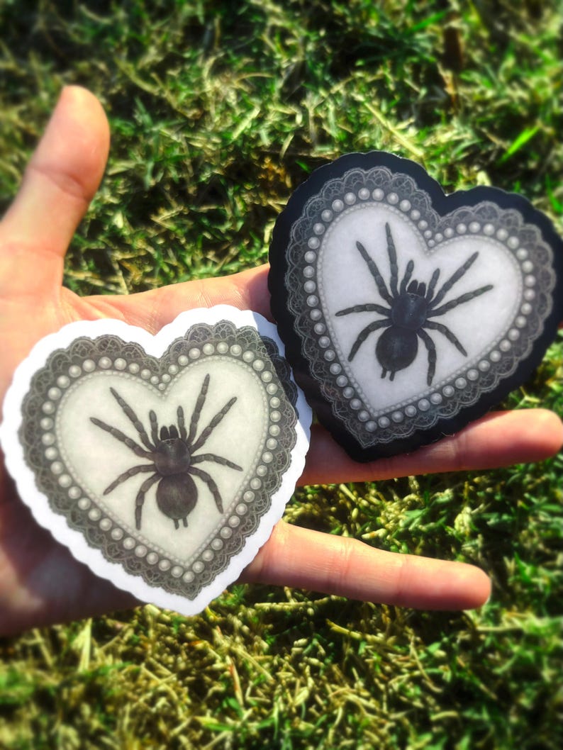 May include: Two heart-shaped stickers with a black spider design. The spiders are on a white background, framed by a grey lace border with pearl accents. The stickers are held against a backdrop of green grass.