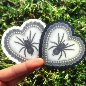 May include: Two heart-shaped stickers with a black spider design. The stickers have a white background with a grey lace border and a pearl-like trim. One sticker is on a white background, the other on a black background.