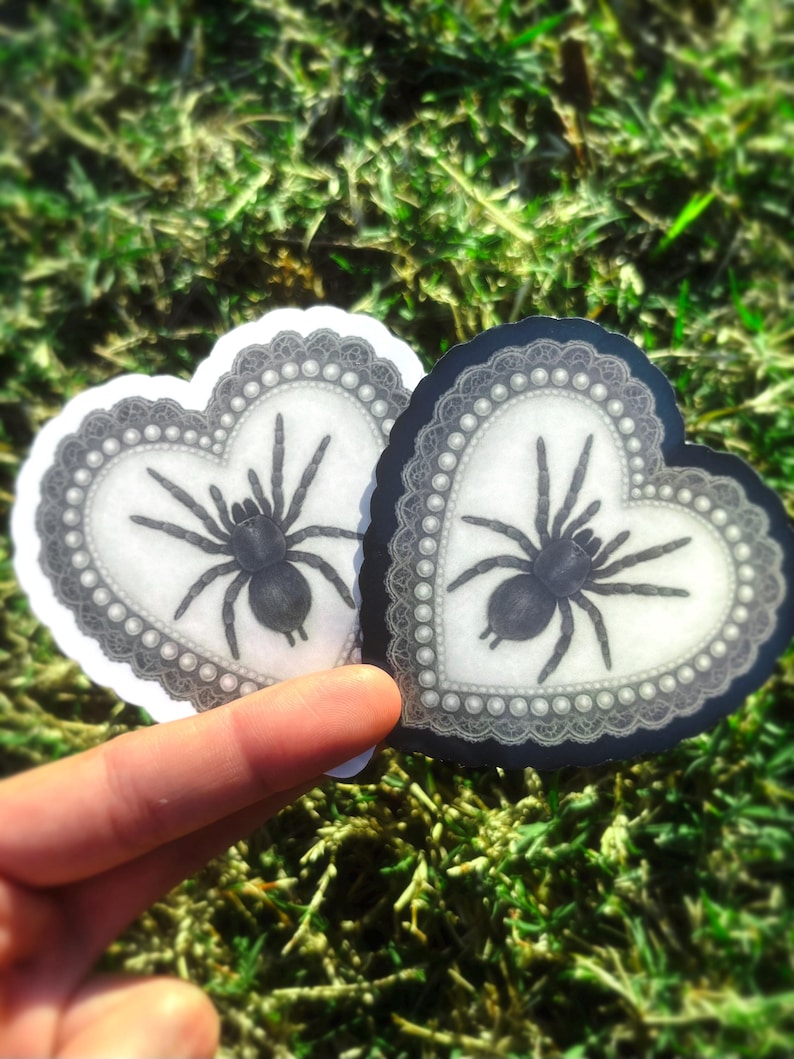 May include: Two heart-shaped stickers with a black spider design. The stickers have a white background with a grey lace border and a pearl-like trim. One sticker is on a white background, the other on a black background.