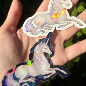 May include: Two unicorn stickers with iridescent accents. The unicorns are depicted in a reclining pose, with flowing manes and tails. The stickers have a holographic effect, with colorful geometric shapes. The background is a blurred green.