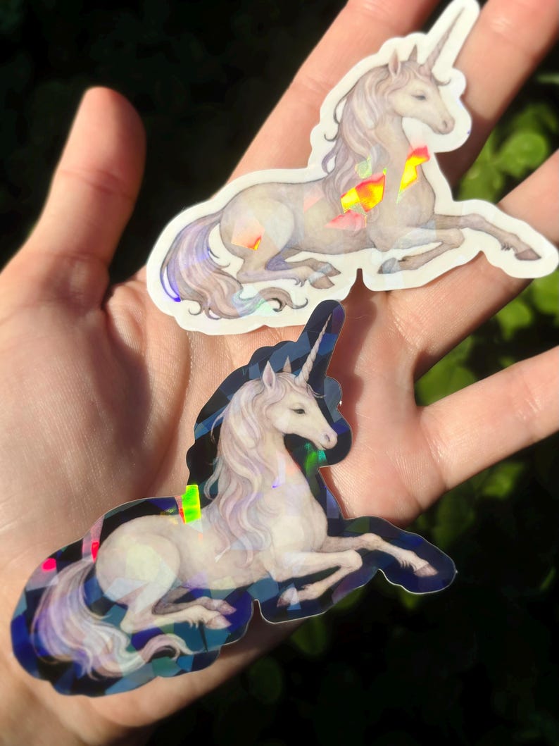May include: Two unicorn stickers with iridescent accents. The unicorns are depicted in a reclining pose, with flowing manes and tails. The stickers have a holographic effect, with colorful geometric shapes. The background is a blurred green.