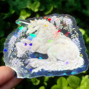 Puede incluir: Una pegatina hologr&aacute;fica que presenta un unicornio blanco con una melena y cola fluidas, rodeado de elementos florales. La pegatina tiene un efecto brillante e iridiscente con acentos coloridos, creando un dise&ntilde;o m&aacute;gico y caprichoso.