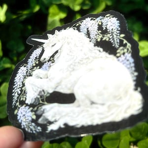 Puede incluir: Adhesivo de un unicornio blanco con borde negro. El unicornio est&aacute; acostado, rodeado de flores moradas claras y hojas verdes. El adhesivo se sostiene frente a un fondo verde borroso.