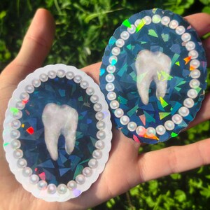 May include: Two oval stickers with a tooth design. The teeth are white, set against a dark blue, geometric background. Each sticker is framed with small, pearl-like beads. One has a white border, the other a holographic border. The stickers are held in a hand.