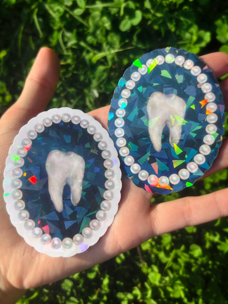 May include: Two oval stickers with a tooth design. The teeth are white, set against a dark blue, geometric background. Each sticker is framed with small, pearl-like beads. One has a white border, the other a holographic border. The stickers are held in a hand.