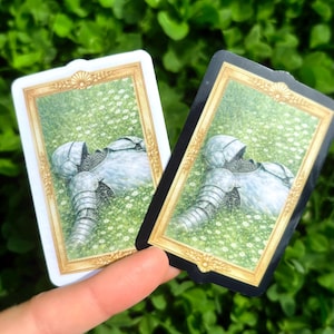 Puede incluir: Dos tarjetas rectangulares con marcos dorados. Cada tarjeta presenta una ilustraci&oacute;n de una armadura tendida en un campo de flores blancas. Una tarjeta tiene un borde blanco, la otra un borde negro. El fondo es verde.