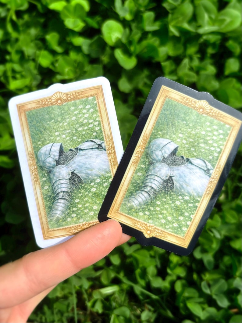 Puede incluir: Dos tarjetas rectangulares con marcos dorados. Cada tarjeta presenta una ilustraci&oacute;n de una armadura tendida en un campo de flores blancas. Una tarjeta tiene un borde blanco, la otra un borde negro. El fondo es verde.