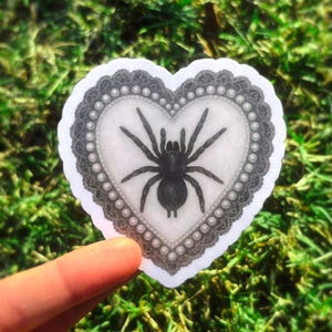 May include: A heart-shaped sticker with a black spider illustration. The spider is in the centre of a white heart, framed by a grey lace border and pearl-like accents. The sticker is held against a backdrop of green grass.