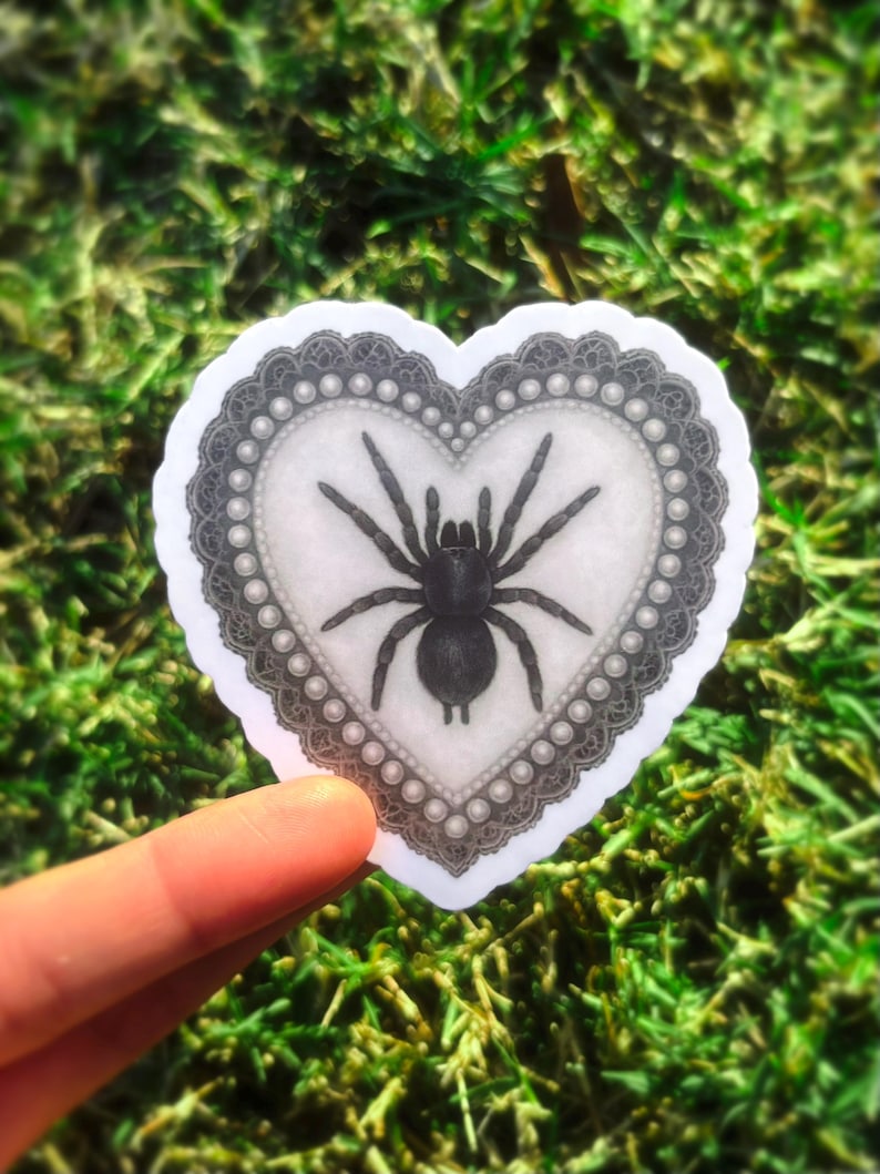 May include: A heart-shaped sticker with a black spider illustration. The spider is in the centre of a white heart, framed by a grey lace border and pearl-like accents. The sticker is held against a backdrop of green grass.
