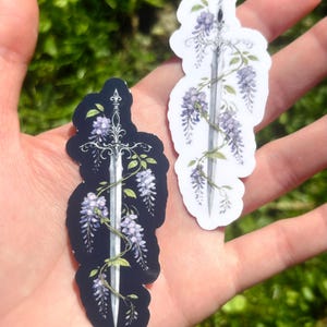 May include: Two stickers featuring a sword design entwined with purple wisteria flowers and green leaves. One sticker has a black background, the other is white. The stickers are held in a hand against a blurred green background.