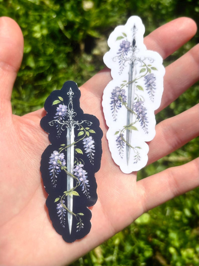 May include: Two stickers featuring a sword design entwined with purple wisteria flowers and green leaves. One sticker has a black background, the other is white. The stickers are held in a hand against a blurred green background.