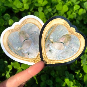 Può includere: Due adesivi a forma di cuore con un dipinto all'interno di una cornice dorata. Un adesivo ha un bordo bianco, l'altro un bordo nero. Il dipinto raffigura una donna in abito bianco. Lo sfondo è verde.