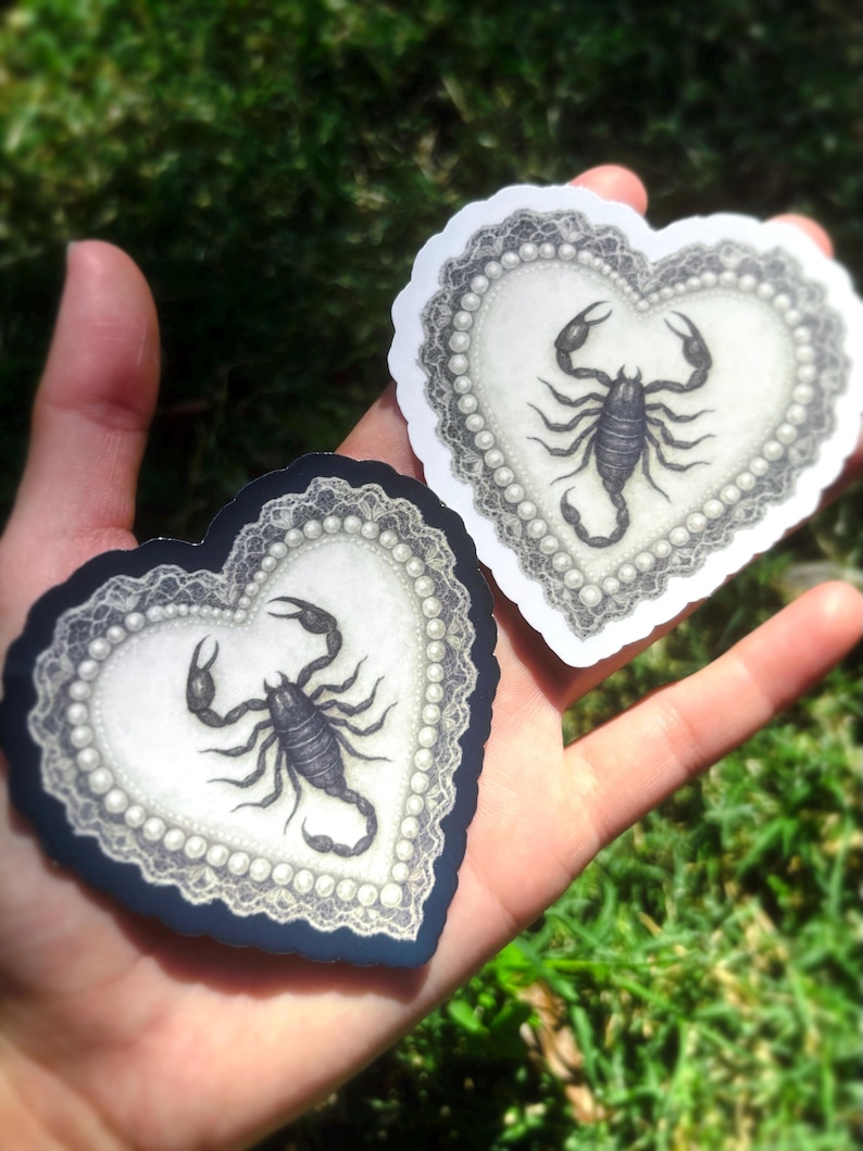 Pu&ograve; includere: Due adesivi a forma di cuore con una dettagliata illustrazione di uno scorpione nero. Ogni adesivo ha un centro bianco con un bordo perlato e un bordo in pizzo decorativo. Un adesivo ha un bordo blu navy, l'altro un bordo bianco.