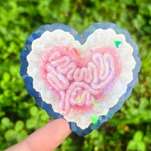 May include: A heart-shaped sticker with a pink brain design and the words "Brain You". The sticker has a white border with a blue and silver iridescent edge. The background is blurred green foliage.