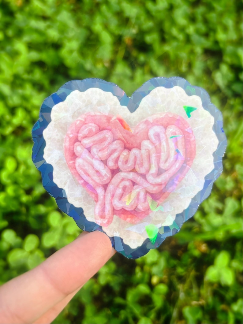 May include: A heart-shaped sticker with a pink brain design and the words "Brain You". The sticker has a white border with a blue and silver iridescent edge. The background is blurred green foliage.
