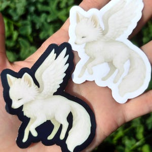 May include: Two die-cut stickers featuring a white winged fox in a leaping pose. One sticker has a solid black background, while the other has a white background. The fox has closed eyes, pointed ears, and large feathered wings extending upward.