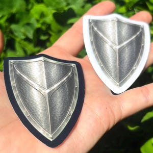 K&ouml;nnte beinhalten: Zwei schildf&ouml;rmige Aufkleber mit metallisch-silbernem Design. Einer hat einen schwarzen Rand, der andere einen wei&szlig;en Rand. Die Schilde haben eine strukturierte Oberfl&auml;che und ein zentrales Designelement. Der Hintergrund ist verschwommenes gr&uuml;nes Laub.
