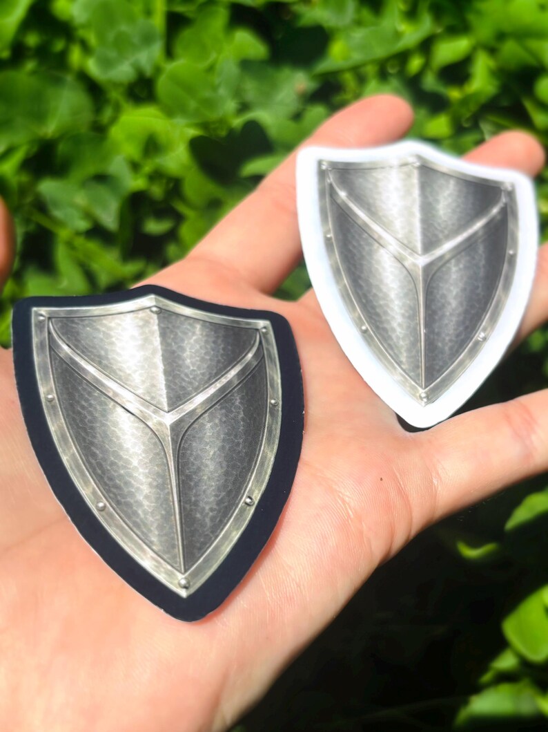 K&ouml;nnte beinhalten: Zwei schildf&ouml;rmige Aufkleber mit metallisch-silbernem Design. Einer hat einen schwarzen Rand, der andere einen wei&szlig;en Rand. Die Schilde haben eine strukturierte Oberfl&auml;che und ein zentrales Designelement. Der Hintergrund ist verschwommenes gr&uuml;nes Laub.
