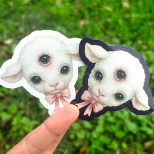 May include: Two stickers featuring a lamb with three eyes and a pink bow. One sticker has a white border, and the other has a black border. The stickers are held up against a blurred green background.