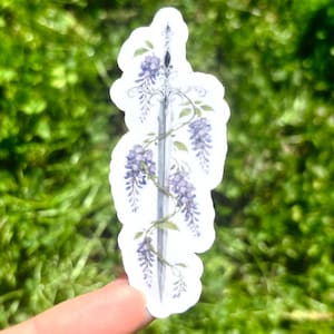 May include: A sticker featuring a detailed illustration of a sword entwined with purple wisteria flowers and green leaves. The sword has a silver blade and ornate hilt. The sticker is on a white background.