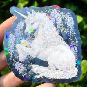 Puede incluir: Una pegatina hologr&aacute;fica con un unicornio blanco con una larga melena y un cuerno en espiral. El unicornio est&aacute; rodeado de flores de glicina moradas sobre un fondo azul oscuro. La pegatina tiene un efecto brillante e iridiscente.