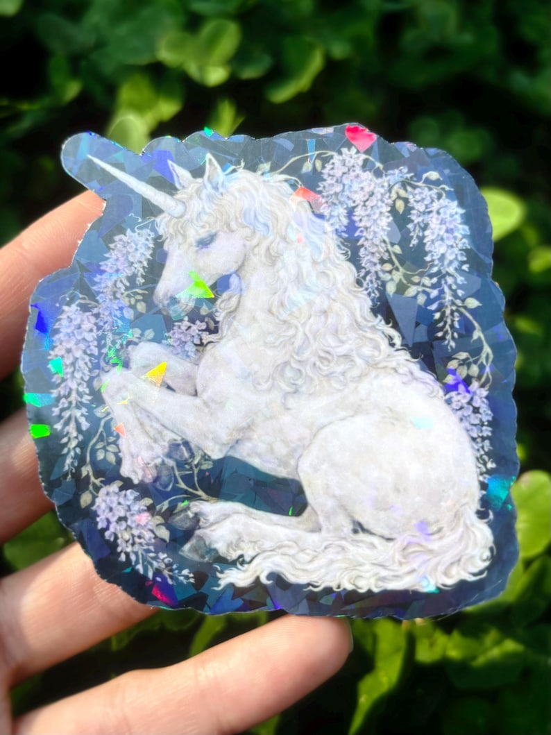 Puede incluir: Una pegatina hologr&aacute;fica con un unicornio blanco con una larga melena y un cuerno en espiral. El unicornio est&aacute; rodeado de flores de glicina moradas sobre un fondo azul oscuro. La pegatina tiene un efecto brillante e iridiscente.