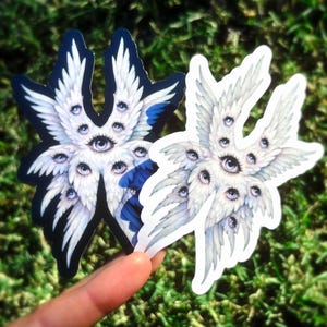 May include: Two stickers featuring a detailed illustration of an angel with multiple eyes and white feathered wings. One sticker has a black border, the other has a white border. The stickers are held against a background of green grass.