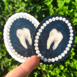 May include: Two round stickers with a tooth design. Each sticker has a dark blue background, a white tooth, and a border of white pearls. One sticker has a white border, the other a black border.