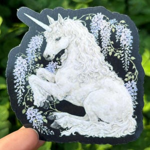 Puede incluir: Una pegatina con un unicornio blanco con un cuerno en espiral, melena rizada y cola larga. El unicornio est&aacute; rodeado de flores de glicina moradas claras y hojas verdes, sobre un fondo negro.